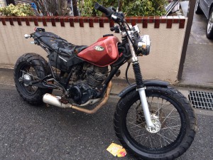 長期放置されたヤマハ TW200の不動車。赤いタンク、カスタムヘッドライト、錆が見られるマフラーと車体フレーム、シートが欠損した状態のオフロードバイク。